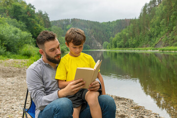 Fototapeta premium Father and young son reading book together, sitting on beach by river or lake