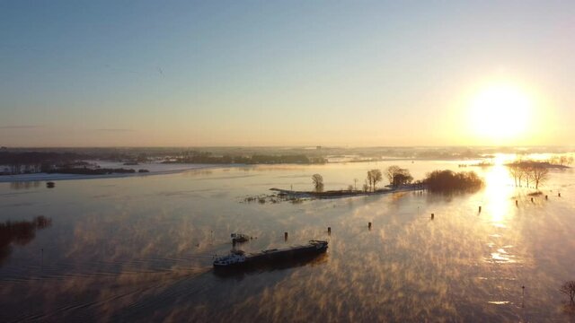 Ship sailing in a foggy winter sunrise over the river Ijssel with ice and snow during a cold day in The Netherlands. Aerial drone point of view.