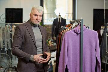 A mature man with gray hair is posing while choosing a turtleneck sweater in a clothing store. A male customer in a wool suit in a boutique.