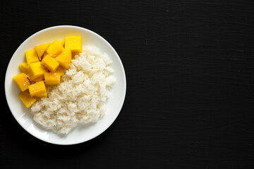Homemade Sticky Rice with Mango and Sesame Seeds on a white plate on a black background, top view. Flat lay, overhead, from above. Space for text.