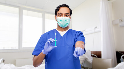 Obraz na płótnie Canvas healthcare, vaccination and medicine concept - indian doctor or male nurse in blue uniform, face protective medical mask and gloves with syringe pointing finger to camera over hospital ward background