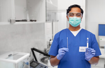 healthcare, profession and medicine concept - indian doctor or male nurse in blue uniform, face mask, goggles and gloves with cotton swab and test tube over laboratory or hospital background