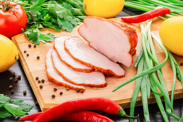 Smoked-cooked pork meat, ham, cut into slices on a cutting wooden board. Nearby are tomatoes, red hot peppers, lemon, parsley, green onions and black pepper. Close-up