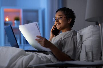 technology, communication and remote job concept - happy smiling young african american woman with laptop computer and papers calling on smartphone in bed at home at night