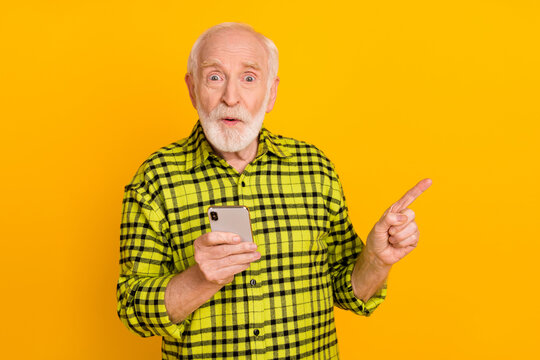 Photo of aged man excited amazed use cellphone indicate finger empty space ad promo advice isolated over yellow color background - Powered by Adobe