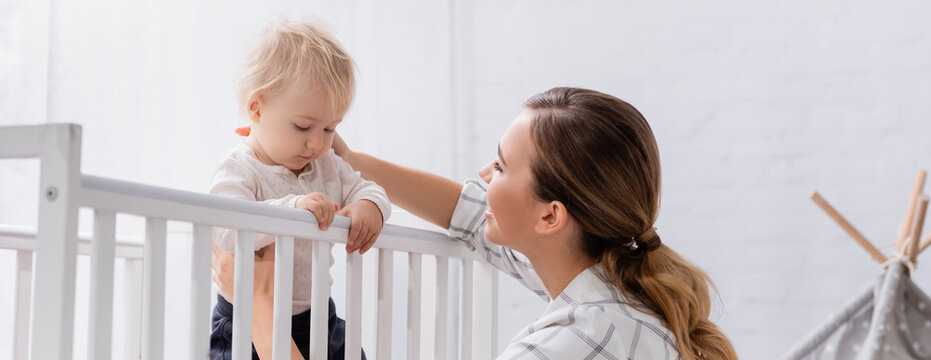 Young Woman Supporting Toddler Son Standing In Crib, Banner