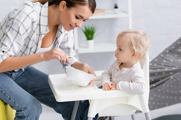 Obraz na płótnie Canvas mother holding bowl near toddler sitting on kids chair