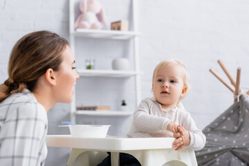 young woman looking at little son sitting on kids chair near bowl, blurred foreground