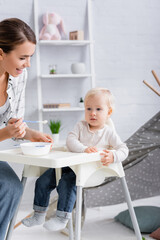 young woman holding spoon with puree near child sitting on kids chair