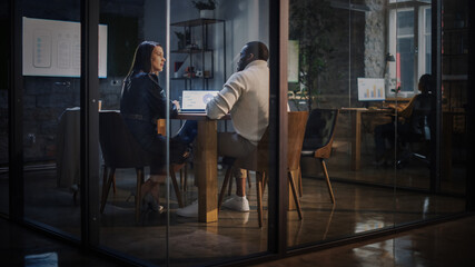 Two Diverse Multiethnic Colleagues Have a Conversation in a Meeting Room Behind Glass Walls in an Agency. Female Creative Director and African American Project Manager Discuss Work on Laptop Computer.