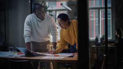 Two Diverse Multiethnic Colleagues Have a Conversation in a Meeting Room Behind Glass Walls in an Agency. African American Creative Director and Female Project Manager Discuss Work on Laptop Computer.