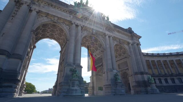 Upwards angle sideways rotating shot of the Cinquantennaire triumphal arc monument in Brussels, Belgium, on warm sunny summer day with blue skies. Belgian flag waving in middle arch. Sun glare above.