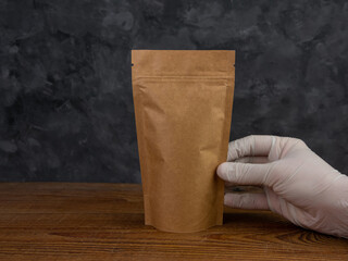 Pouch bag mockup. Hand in protective gloves holds brown kraft paper pack with coffee beans wooden background. Packaging with clasp template food delivery tea leaves weight product. Shop sale ad design