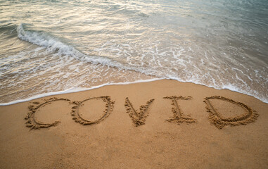 Handwriting COVID on sand and foam wave on beach. Coronavirus concept.