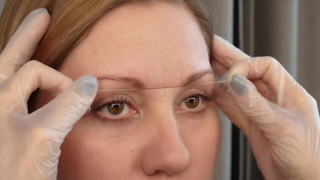 Professional styling of client face and stylist's hands working with special thread to mark new contour of brows. Correction and shaping procedures of female eyebrows in beauty salon