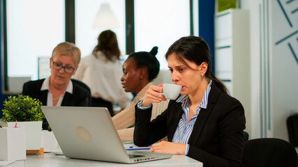 Concentrated manager typing on laptop sitting at desk in start up business office drinking coffee while diverse colleagues working in background. Multiethnic coworkers planning new financial project.