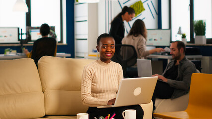 Portrait of african woman typing on laptop looking at camera smiling while diverse team working in background. Multiethnic coworkers talking about startup financial company in modern business office
