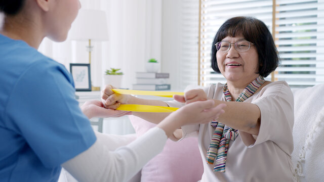 Young older senior asia citizen sit on sofa couch with in-home nursing care, assisted living, scrubs nurse doctor use resistance band exercise for senior patient in physiotherapy treatment at home. - Powered by Adobe