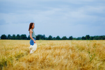 A thin girl with a hat in her hand is spinning in the middle of a wheat field against the background of the sky with clouds. Rural summer landscape.