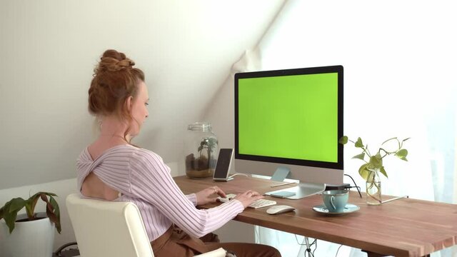 Young woman sitting in front of a monitor with green screen chroma key. Woman is working at home office in front of a computer with coffee cup. Concept for Website, Application or Business. 4K.
