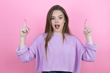 Young pretty woman with long hair standing over isolated pink background amazed and surprised looking at the camera and pointing up with fingers and raised arms