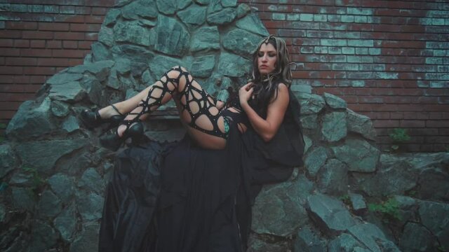 Fantasy woman Medusa gorgon Greek goddess. Black gothic carnival costume. Girl Fashion model girl sitting on stone throne, posing. On head are golden snakes hair, scary makeup. Dark queen lady witch