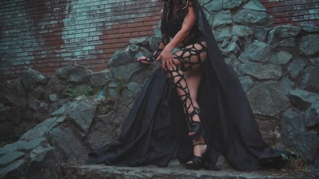 Fantasy woman Medusa gorgon Greek goddess. Black gothic carnival costume. Girl Fashion model girl sitting on stone throne, posing. On head are golden snakes hair, scary makeup. Dark queen lady witch