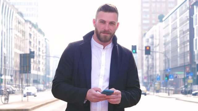 Slow motion young handsome bearded businesslike adult man outdoor backlight using smartphone looking camera smiling positive
