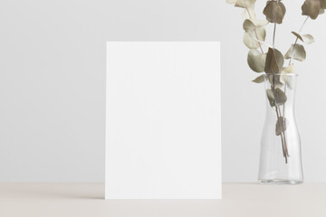 White invitation card mockup with an eucalyptus on the beige table. 5x7 ratio, similar to A6, A5.