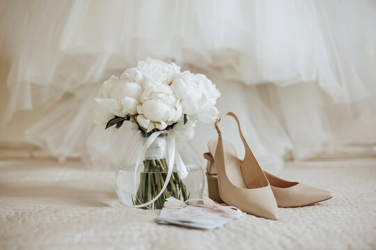 Wedding bouquet of peonies flowers in a vase stands on the bed of the newlyweds with invitations and shoes on the background of the bride dresses - Powered by Adobe