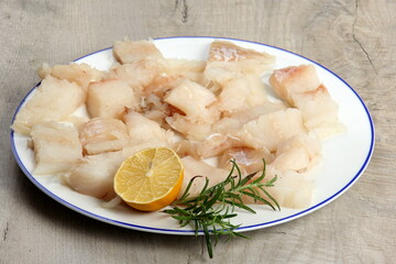 Pieced of sea fish filet on a white plate decorated with lemon and rosemary, white raw fish on a plate, gray wooden background