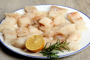 Pieced of sea fish filet on a white plate decorated with lemon and rosemary, white raw fish on a plate, gray wooden background