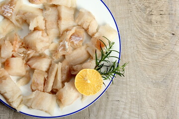 Pieced of sea fish filet on a white plate decorated with lemon and rosemary, white raw fish on a plate, gray wooden background