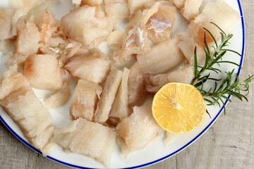 Pieced of sea fish filet on a white plate decorated with lemon and rosemary, white raw fish on a plate, gray wooden background