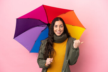 Young woman holding an umbrella isolated on pink background pointing to the side to present a product