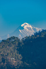 A view of the Trishul mountain peak on the Himalayan range with a hill with trees in the foreground