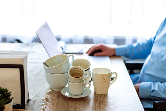 Bad Lifestyle Caffeine Addicted Concept. Many Cups Of Coffee On The Desk. A Man Working From Home On Laptop As The Background. Drinks Too Much Coffee Caffeine Addiction Anxious And Crazy In Maniac. 
