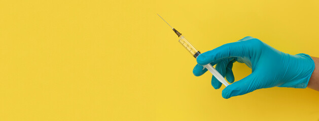 Doctor's hand in blue medical (surgical) gloves holding a plastic syringe on yellow background.  Banner size with space for text.