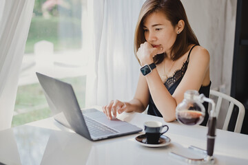 Working from home concept, an Asian woman is using her laptop to check email and work via the internet with a coffee next to her. Have a comfortable mood