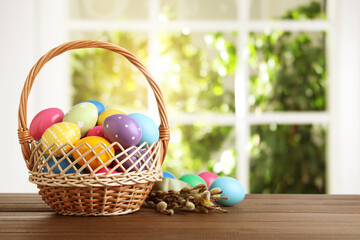 Colorful Easter eggs in wicker basket and willow branches on wooden table, space for text