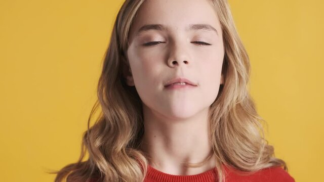 Close up beautiful blond teenage girl with wavy hair blowing bubble from gum on camera and happily smiling over yellow background. Funny girl blowing candy bubble