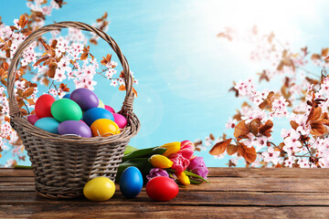 Colorful Easter eggs in wicker basket and tulips on wooden table outdoors, space for text
