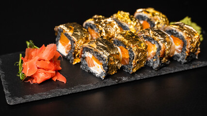 Custom sushi roll with nori, cuttlefish ink, fresh salmon, cream cheese, pepper pumpkin, eel, unagi sauce and edible gold leaf with red ginger, wasabi on a square plate on black table and background.