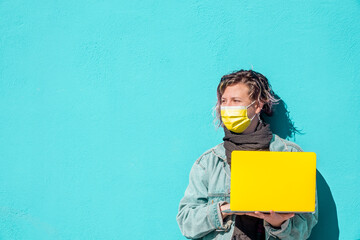 Student with a yellow computer and yellow protection mask on the turqoise wall background