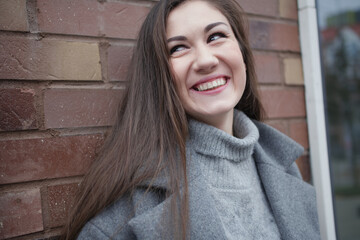 Portrait of a charming cheerful girl on the background of a brick wall in the street. The girl looks to the side and laughs cheerfully. A beautiful girl in a coat walks around the city