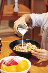 Cropped image of woman pouring glass of milk in bowl of muesli with dry berries or raisins