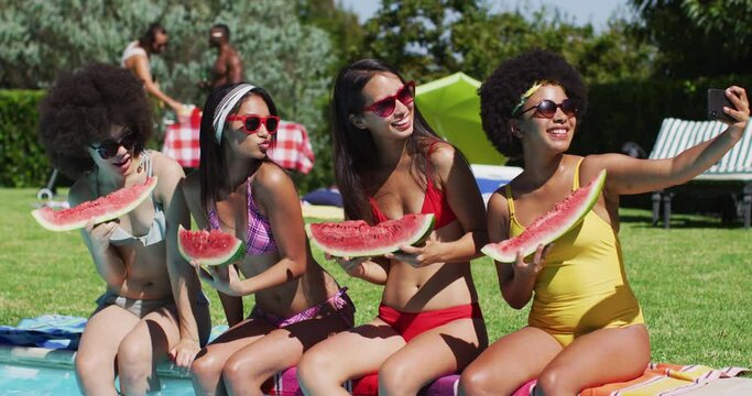 Group of diverse girls holding watermelon taking a selfie together on smartphone while sitting by th