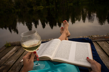 girl on the riverbank reading a book and drinking wine in a clear glass, selective focus
