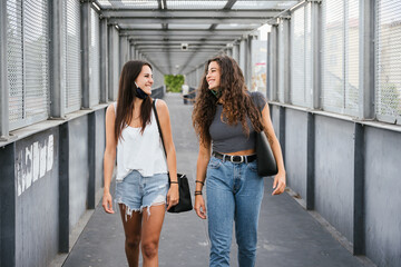 Two beautiful young women walk outdoors around city with face masks down during the Covid-19 Coronavirus pandemic - Millennials girlfriends have fun together talking and joking on a weekend break