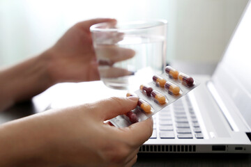Obraz premium Female hands with pills and water glass on laptop keyboard. Woman taking medication in capsules during work
